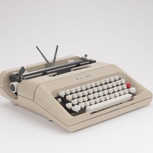 以下が含まれることがあります： QWERTYキーボード、白い円形のキー、右側に赤いキーが1つ、黒いキャリッジを備えたベージュのポータブルタイプライター。給紙機構近くのトップケースにはErika 15というブランド名が印字されています。
