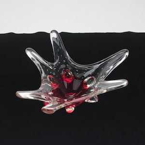 May include: A clear glass bowl with an irregular, star-like shape featuring multiple pointed arms. The center of the bowl contains deep red glass accents and small air bubbles embedded within the clear material, creating a textured appearance.