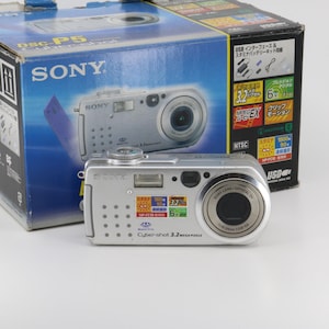 Könnte beinhalten: Eine silberne Sony Cyber-shot DSC-P5 Digitalkamera mit einer Auflösung von 3,2 Megapixel und einem 3-fach optischen Zoomobjektiv. Die Kamera hat ein silbernes Gehäuse mit schwarzen Knöpfen und einem schwarzen Objektiv. Die Kamera steht auf einer weißen Oberfläche.
