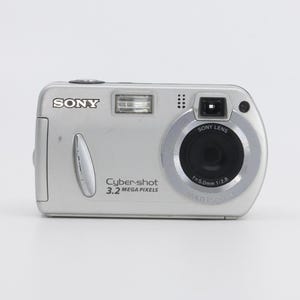 Könnte beinhalten: Eine silberne Sony Cyber-shot Digitalkamera mit einer Auflösung von 3,2 Megapixel und einem 5,0 mm Objektiv. Die Kamera hat ein schwarzes Objektiv und ein silbernes Gehäuse mit dem aufgedruckten Text "SONY" und "Cyber-shot".