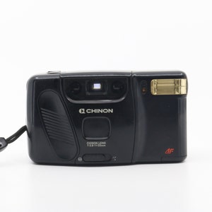 Op de afbeelding: Een zwarte compactfilmcamera met een zilveren Chinon-logo en een gouden flitser. De camera heeft een Chinon-lens met een 1:3.9 f=35mm lens.