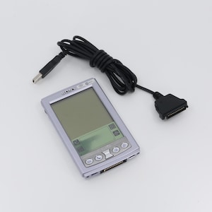 Könnte beinhalten: Ein silberner Sony Clie PDA mit einem schwarzen USB-Kabel und einem schwarzen Stecker. Der PDA hat einen kleinen Bildschirm und mehrere Tasten.