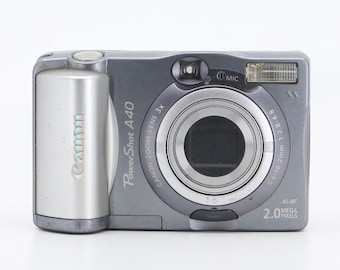 Pode incluir: Uma câmera digital Canon PowerShot A40 prateada com uma lente zoom 3x e 2,0 megapixels. A câmera tem uma lente preta e um corpo prateado.