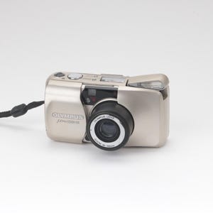 May include: A champagne-coloured Olympus Mju Zoom 105 point-and-shoot film camera with a retractable lens cover, a black lens barrel labelled 38-105mm, a built-in flash, and a black wrist strap attached to the side of the compact metal body.