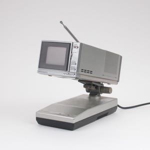 Könnte beinhalten: Ein silber-schwarzer tragbarer Panasonic Mikro-Farbfernseher mit kleinem Bildschirm, ausziehbarer Antenne und schwenkbarem Sockel. Das Gerät verfügt über Bedientasten an der Vorderseite und ein Lautsprechergitter an der Seite des Gehäuses.