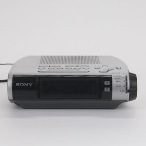 May include: A silver and black Sony digital alarm clock radio featuring a central display screen, a speaker grille on top, and a side dial for alarm mode settings labeled OFF, RADIO, BUZZER, and A+B. The front panel includes buttons for A and B.