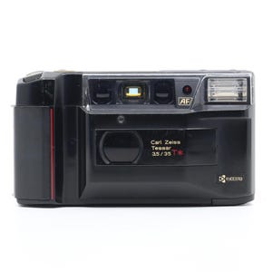 May include: A black Kyocera camera with a Carl Zeiss T* lens. The camera has a built-in flash and an autofocus system.