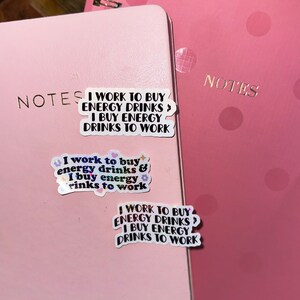 K&ouml;nnte beinhalten: Zwei rosa Notizb&uuml;cher mit dem Wort "NOTES" in goldfarbenen Buchstaben. Auf dem Notizbuch befinden sich drei Aufkleber, die jeweils den Text "I WORK TO BUY ENERGY DRINKS? I BUY ENERGY DRINKS TO WORK" tragen. Ein Aufkleber hat florale Akzente.