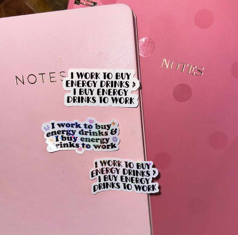K&ouml;nnte beinhalten: Zwei rosa Notizb&uuml;cher mit dem Wort "NOTES" in goldfarbenen Buchstaben. Auf dem Notizbuch befinden sich drei Aufkleber, die jeweils den Text "I WORK TO BUY ENERGY DRINKS? I BUY ENERGY DRINKS TO WORK" tragen. Ein Aufkleber hat florale Akzente.