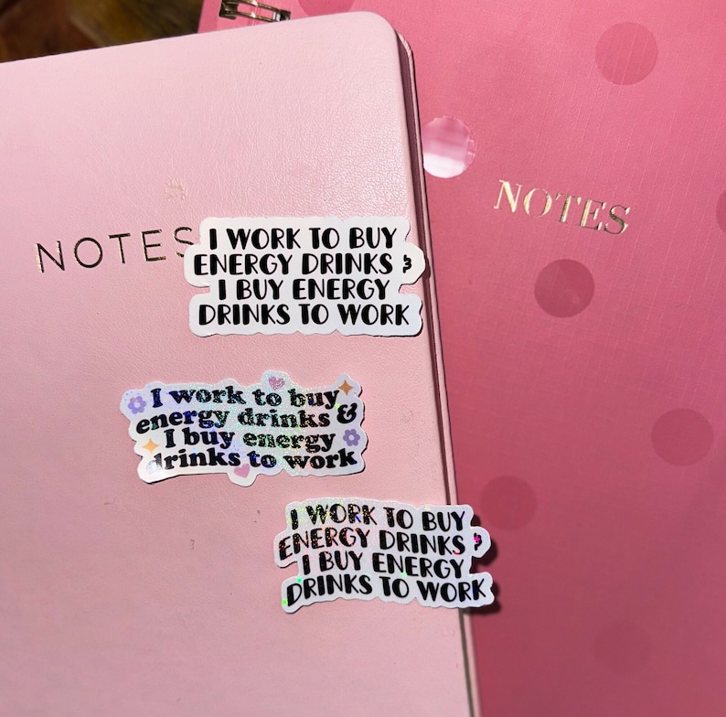 K&ouml;nnte beinhalten: Zwei rosa Notizb&uuml;cher, eines mit goldfarbenem Schriftzug "NOTES". Auf den Notizb&uuml;chern befinden sich drei Aufkleber. Die Aufkleber tragen den Text "I WORK TO BUY ENERGY DRINKS & I BUY ENERGY DRINKS TO WORK". Ein Aufkleber hat florale Akzente.