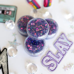 May include: Four small, clear plastic jars filled with purple, pink, and teal glitter are stacked on a white surface. The glitter is a mix of fine and chunky particles. A purple patch with the word "SLAY" is visible.