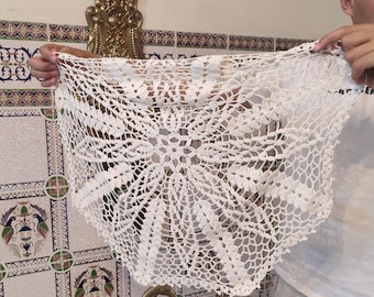 May include: A white, open-weave crochet shawl with a floral pattern. The shawl is held up, showcasing its intricate design and delicate texture. The background includes decorative wall tiles and a bronze horse statue.
