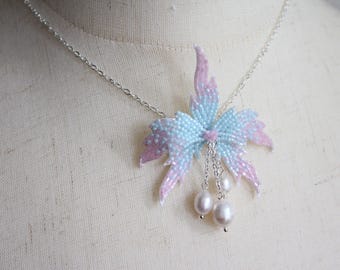May include: A beaded necklace featuring a floral pendant with a gradient of light blue and pink. The pendant is adorned with dangling white pearls and hangs from a delicate silver chain. The necklace is displayed on a mannequin.