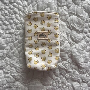 May include: A small, rectangular pouch with a cream-colored background and a repeating pattern of yellow duck illustrations. A small, rectangular patch with a mountain and tree design is sewn on the front. The pouch is likely made of fabric.