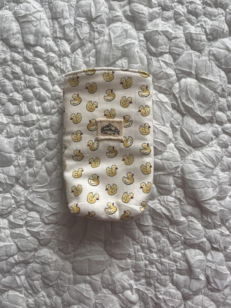 May include: A small, rectangular pouch with a cream-colored background and a repeating pattern of yellow duck illustrations. A small, rectangular patch with a mountain and tree design is sewn on the front. The pouch is likely made of fabric.