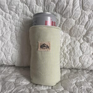 May include: A light green gingham can cooler with a small embroidered patch featuring a mountain design. The cooler holds a silver can with the word "BUBBL" in pink. The background is a white, textured fabric.