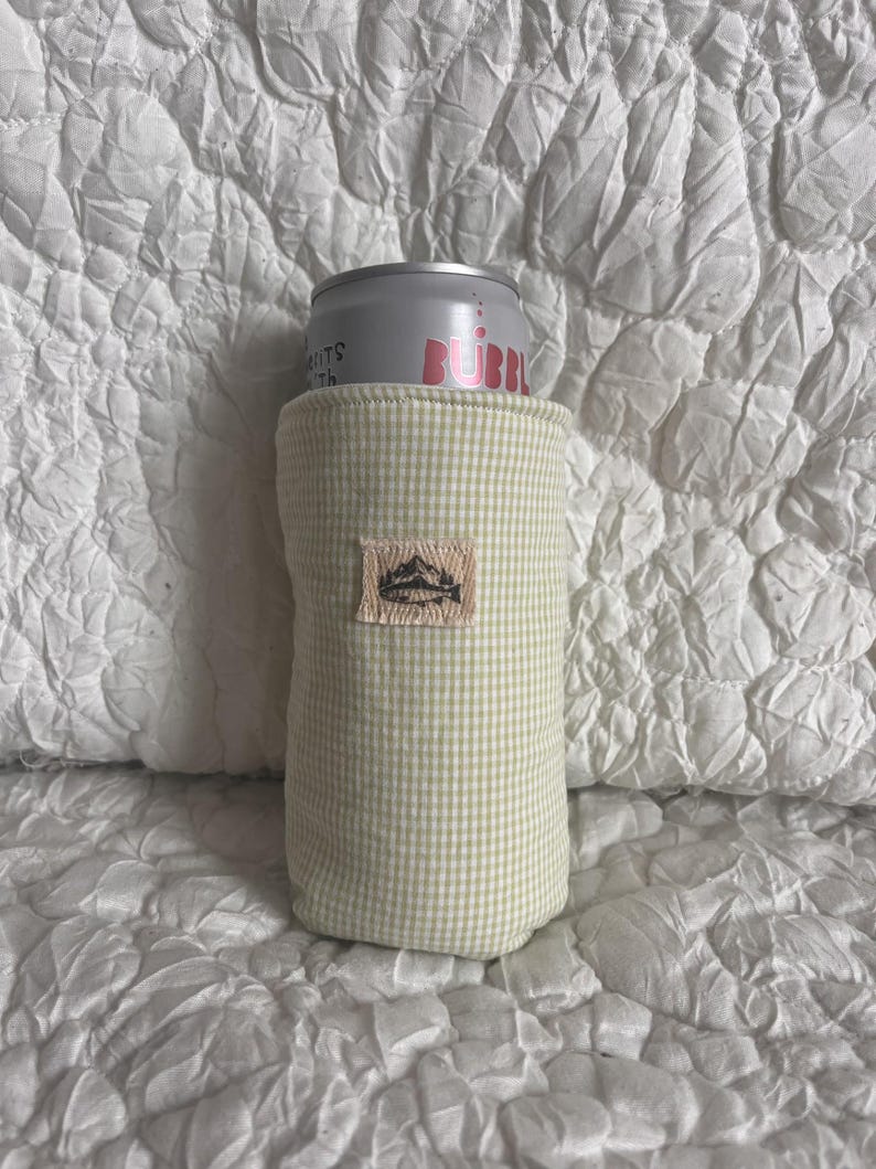 May include: A light green gingham can cooler with a small embroidered patch featuring a mountain design. The cooler holds a silver can with the word "BUBBL" in pink. The background is a white, textured fabric.