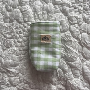 May include: A small, green and white gingham patterned pouch. The pouch has a rectangular patch with a mountain and tree design. The pouch is made of fabric and is likely used for carrying small items.