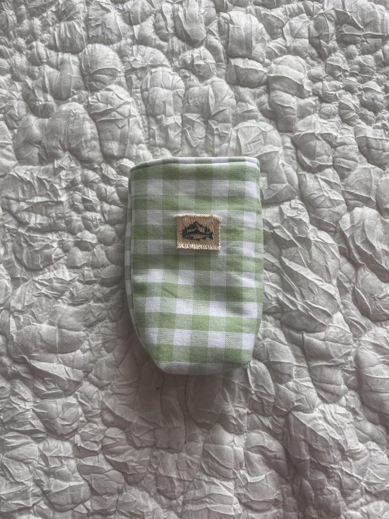 May include: A small, green and white gingham patterned pouch. The pouch has a rectangular patch with a mountain and tree design. The pouch is made of fabric and is likely used for carrying small items.