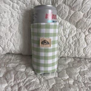 May include: A green and white gingham can cooler with a small rectangular patch featuring a mountain and lake design. A silver can of Bubbl sparkling water is inside the cooler.