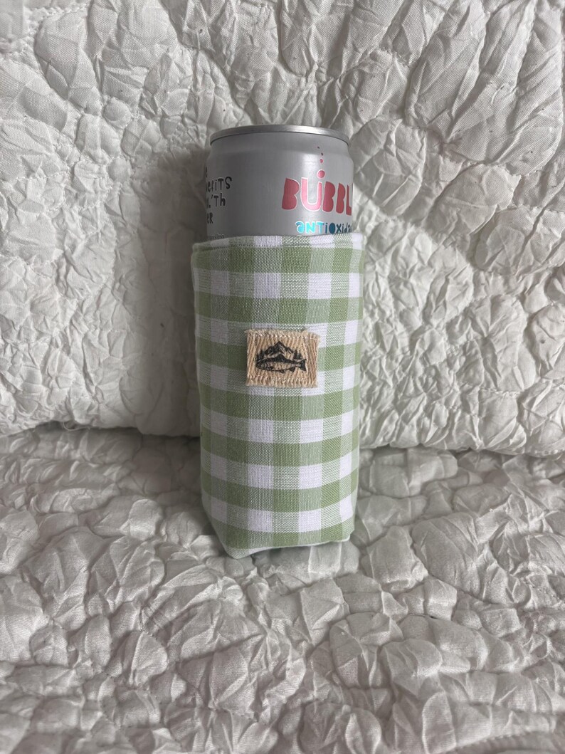 May include: A green and white gingham can cooler with a small rectangular patch featuring a mountain and lake design. A silver can of Bubbl sparkling water is inside the cooler.