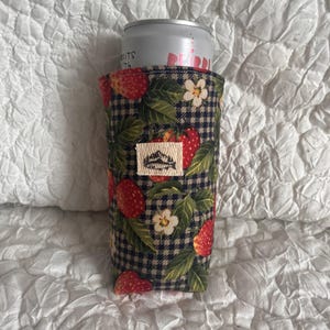 May include: A fabric can cooler featuring a strawberry and floral pattern on a navy and white checkered background. The cooler has a small rectangular tag with a mountain logo. A silver can is inside the cooler.