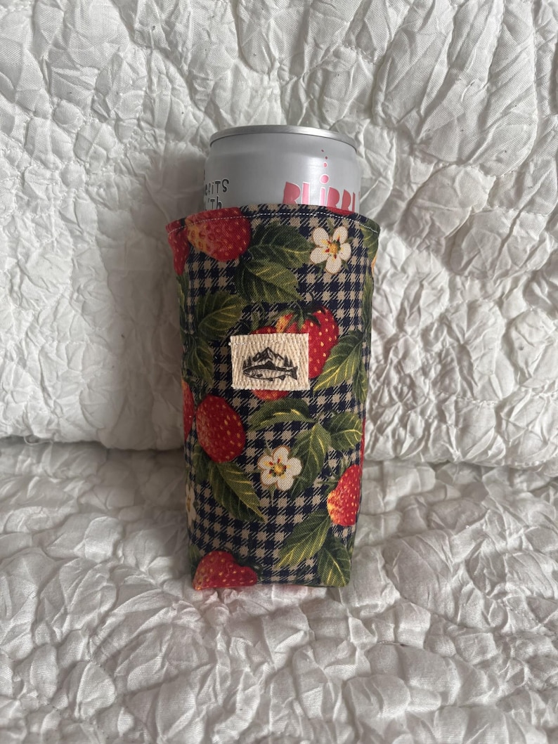 May include: A fabric can cooler featuring a strawberry and floral pattern on a navy and white checkered background. The cooler has a small rectangular tag with a mountain logo. A silver can is inside the cooler.