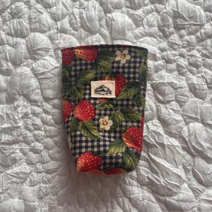 May include: A fabric coffee cup sleeve featuring a strawberry and leaf pattern over a black and white checkered background. The sleeve has a small rectangular tag with a mountain design. The sleeve is designed to fit around a standard coffee cup.