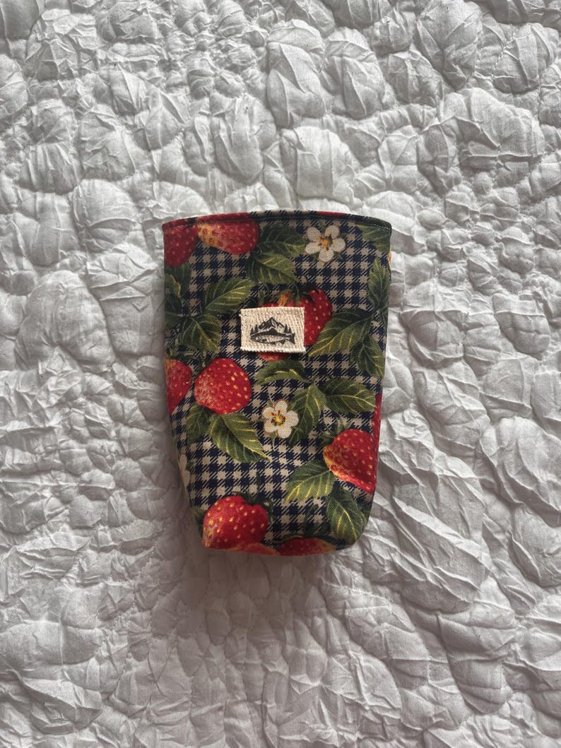 May include: A fabric coffee cup sleeve featuring a strawberry and leaf pattern over a black and white checkered background. The sleeve has a small rectangular tag with a mountain design. The sleeve is designed to fit around a standard coffee cup.