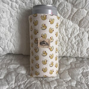 May include: A white fabric can cooler featuring a repeating pattern of yellow duck illustrations. A small rectangular tag with a mountain design is sewn on the cooler. A silver can is inside the cooler.