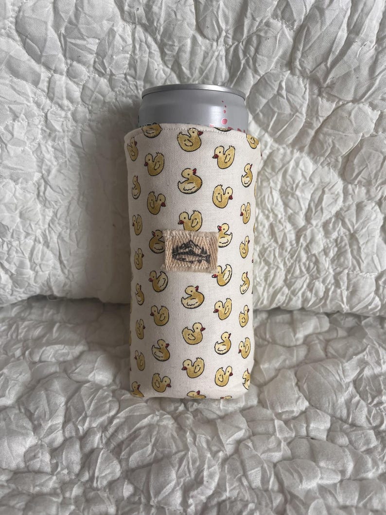 May include: A white fabric can cooler featuring a repeating pattern of yellow duck illustrations. A small rectangular tag with a mountain design is sewn on the cooler. A silver can is inside the cooler.