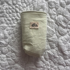 May include: A light green and white gingham patterned fabric container with a small rectangular patch featuring a mountain and lake design. The container is cylindrical and appears to be made of fabric. The background is a textured, light gray surface.