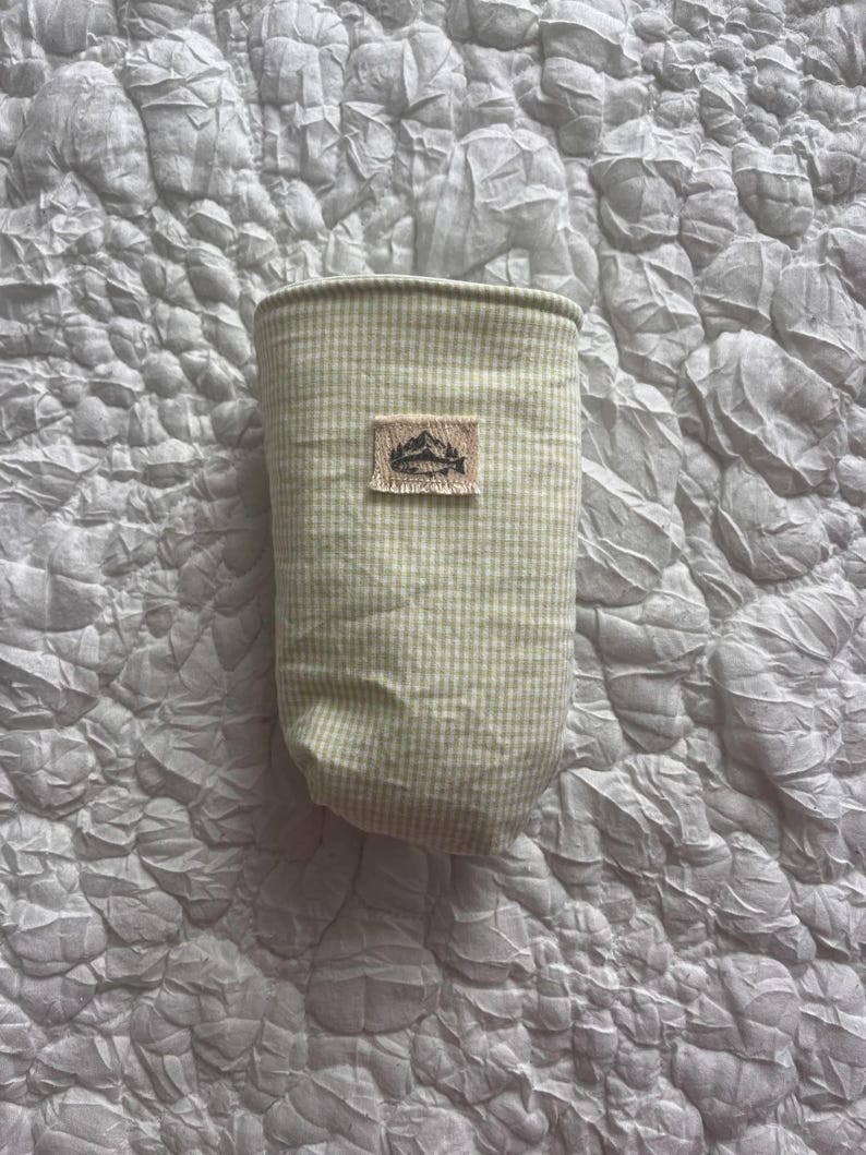 May include: A light green and white gingham patterned fabric container with a small rectangular patch featuring a mountain and lake design. The container is cylindrical and appears to be made of fabric. The background is a textured, light gray surface.