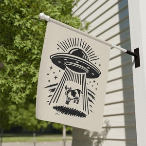 May include: A beige flag featuring a black and white illustration of a UFO beaming up a cow. The design includes a flying saucer, a cow, stars, and light beams. The flag is mounted on a white pole.