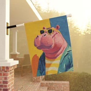 Könnte beinhalten: Dekorative Outdoor-Flagge mit einem rosa Nilpferd, das eine Sonnenbrille und eine blaue Jacke trägt. Das Nilpferd ist vor einem gelb-blauen Hintergrund platziert. Die Flagge ist an einer weißen Stange befestigt und an einer Ziegelstruktur montiert.