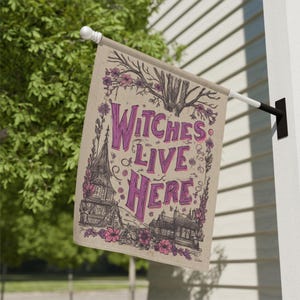 Puede incluir: Una bandera beige para exteriores con el texto "Witches Live Here" en morado. La bandera presenta ilustraciones de casas, flores y ramas de árboles. La bandera está colgada de un poste blanco.