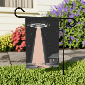 May include: A black and white garden flag featuring a UFO hovering over a house. The UFO is emitting a beam of light. The flag is displayed on a black metal stand.