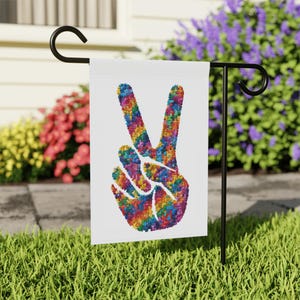 May include: A white garden flag featuring a peace sign formed from a mosaic of colourful puzzle pieces. The flag is mounted on a black metal stand, set in a garden with flowers in the background.