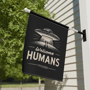 May include: Black flag featuring a retro design of a flying saucer and two figures, with the text "Welcome HUMANS" in white. The flag is mounted on a white wall.