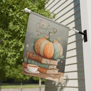 May include: A decorative garden flag features a watercolor illustration of a pumpkin atop a stack of books, with a teacup and saucer. The pumpkin is in shades of orange, green, and tan, with floral vines. The books are brown and green.