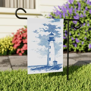 May include: A white garden flag featuring a blue and white illustration of a lighthouse under a cloudy sky. The flag is displayed on a black metal stand in a grassy yard, with colorful flowers in the background.