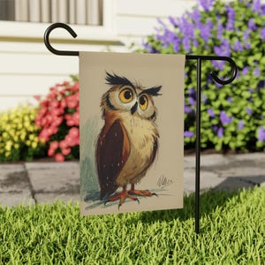 May include: A garden flag featuring a detailed illustration of an owl. The owl has large yellow eyes, brown and beige feathers, and is perched on a branch. The flag is displayed on a black metal stand in a garden setting.