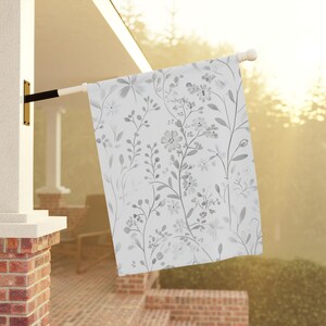 Puede incluir: Una bandera blanca con un patrón floral gris. La bandera está colgada de un poste.