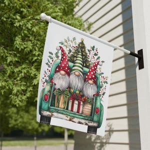 May include: A white garden flag features three gnomes in a green truck bed filled with gifts and greenery. The gnomes wear red and green hats and have long white beards. The truck bed has red circular lights on the sides.