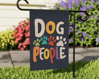 Peut inclure: Un drapeau de jardin bleu marine avec les mots "DOG PEOPLE" en lettres colorées et usées. Le drapeau présente des empreintes de pattes de différentes couleurs. Le drapeau est affiché sur un support en métal noir dans un jardin herbeux.