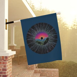 Puede incluir: Una bandera azul marino con un diseño circular que presenta un paisaje montañoso dentro de una vibrante puesta de sol y un centro negro. El diseño está rodeado de líneas radiales. La bandera está montada en un mástil blanco.