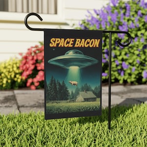 May include: A black garden flag with the words "SPACE BACON" in yellow. The flag features a retro-style illustration of a UFO beaming up a pig over a rural landscape with a house and trees.