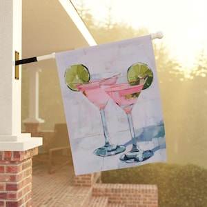 May include: A decorative outdoor flag featuring a painting of two pink cocktails in martini glasses, each garnished with a lime slice. The background is a light, abstract design with brushstrokes, suggesting a sunny day.