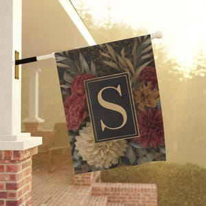 Puede incluir: Bandera decorativa para exteriores con un diseño floral de dalias burdeos, crema y doradas. La bandera tiene un rectángulo negro con la letra "S" dorada. Montada en un mástil blanco.