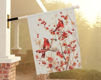 May include: A decorative outdoor flag featuring a watercolor painting of three red cardinals perched on a branch with red berries. The flag is white with a pole and is hanging outside a house.